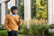 © Liubomir - Confident man standing outside modern office building, holding phone. Wearing casual orange sweater, enjoying sunny day. Reflection of trees seen on glass windows, creating calm serene atmosphere.