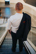 © qunica.com - A rear view of a businessman in a white shirt and jacket descending an escalator in a modern building during the daytime.