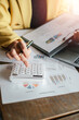 © Satori Studio - Close-up of a businesswoman using a calculator and reviewing financial charts and graphs in an office environment.
