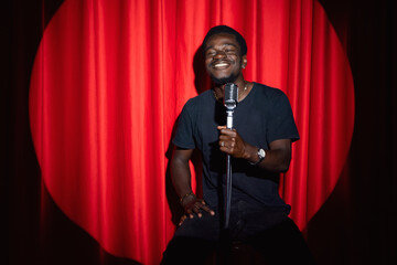 Wall Mural - Happy young african american man comic performing standup monologue on stage with red curtains. Concept comedian show open mike