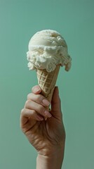 A close-up of a hand holding a single scoop of vanilla ice cream in a waffle cone against a mint green background.