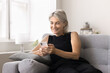 © fizkes - Mature woman using smartphone seated on couch at home, managing tasks, scheduling activities, enjoy on-line shopping, editing images, or creating content for social media using new mobile application