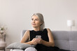 © fizkes - Pensive mature woman sits on sofa holding smartphone looking away thinking, wait for call from friend spend time alone in living room. Modern gadget usage for communication, making order or phonecall