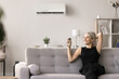 © fizkes - Satisfied middle-aged female resting seated on sofa holds remote controller adjusting, switch-on air conditioner, cooling living room on hot summer day, enjoy comfort and convenient life at smart home