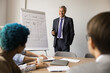 © fizkes - Older 60s smiling man in suit presenting new project to group of people listening intently seated around conference table, take part in business meeting, training session, strategic planning workshop