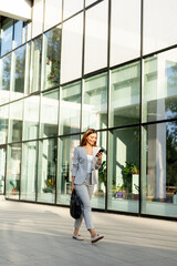 Wall Mural - Business woman strolls confidently outdoors in smart attire, checking her messages against the backdrop of a modern glass building on a sunny day