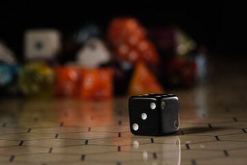 a black six-sided D6 dice with white dots, showing three dots, sitting on a hexagonal tabletop gaming board with a variety of different types of dice in the background. 