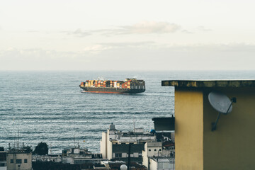 A telephoto shot of a large container ship sails on the open ocean near the coast. The vessel is loaded with multiple colorful cargo containers captured from a vantage point overlooking a coastal city