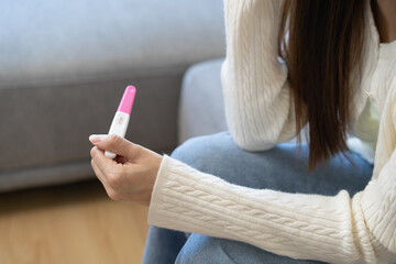 Sad, Disappointed motherhood asian young woman hand holding pregnancy test, upset ovulation problem, anxiety after result at positive sitting on couch at home. Unwanted Pregnant, contraception concept