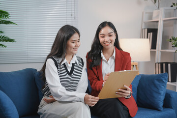 Two smiling businesswomen are having a productive meeting while sitting on a comfortable blue sofa and reviewing information from a clipboard