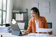 © amnaj - Asian businesswoman is smiling while working on a tablet and reviewing paperwork at her desk