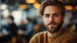 © aicandy - A bearded man with a warm smile in a cozy sweater sitting in a bustling cafe, with a blurred background of people, creating a friendly and welcoming atmosphere