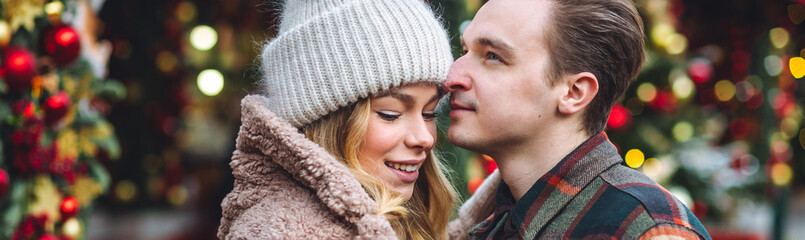 Wall Mural - Beautiful young loving couple embracing, kissing, enjoying winter vacation, cold snowy day on a street city Christmas market outdoors. Warm clothes, knitted sweater, mittens. Cozy atmosphere banner