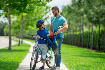 Canvas Print - Father helping son get ready for school. Father and son cycling on bike on summer day. Father support child. Fathers love. Sporty kids. Cute boy with dad cycling in summer park outdoor.