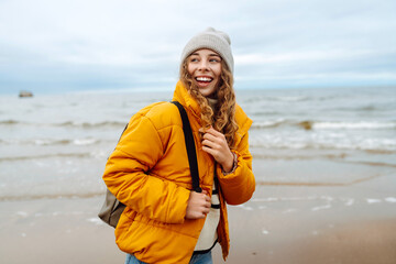 Wall Mural - Young woman in a yellow jacket smiles on the beach near the ocean, enjoying a windy day with gray skies. Calmness and tranquility. Travel, tourism concept.