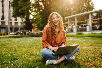 Wall Mural - A young woman enjoying a sunny afternoon working on her laptop in a park setting surrounded by greenery and urban views. Freelancer, Online education.