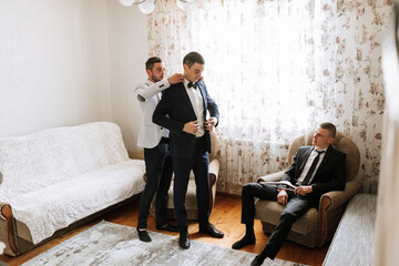 Three men are in a living room, one of them is getting ready to wear a tie. The other two men are sitting on chairs, one of them reading a book. Scene is casual and relaxed