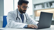 © halo - A male doctor in a white coat is using a laptop computer while sitting at a desk, working on digital medical documents and data online.