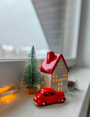 A candlestick house, a red retro car and a Christmas tree decorated with a garland stand on the windowsill. Window sill decoration for the New Year