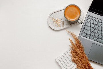 Cozy home office workspace with laptop, cup of coffee, tray and pampas with copy space on white wooden table, top view. Office workplace background, aesthetic style