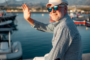 happy senior man in casual shirt and hat sitting at the sea port greeting friends. relaxed elderly b