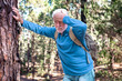© luciano - Senior bearded man feeling bad while walking in the mountain woods. Elderly man with backpack touching his head leaning against a tree trunk to catch his breath
