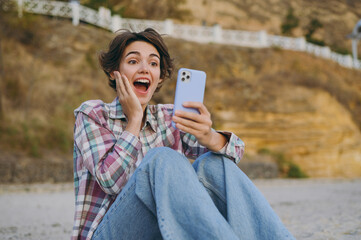 Wall Mural - Young surprised shocked happy woman wears shirt casual clothes hold in hand use mobile cell phone sitting rest on sea ocean sand shore beach outdoor seaside in summer day free time. Lifestyle concept.