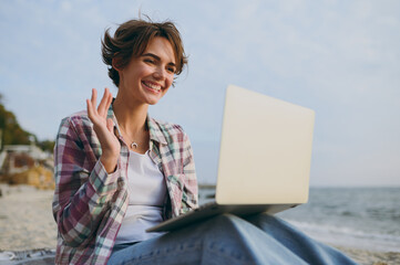 Wall Mural - Side view young IT woman wear shirt casual clothes hold use work on laptop pc computer waving hand sits rest on sea ocean sand shore beach outdoor seaside in summer day free time. Lifestyle concept.