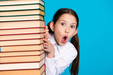 Wall Mural - Portrait of little schoolchild peek pile stack book wear uniform isolated on blue color background