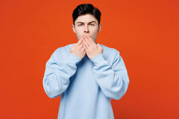 Poster - Young sad shocked surprised Caucasian man he wears blue sweatshirt casual clothes cover mouth with hand looking camera isolated on plain red orange color background studio portrait. Lifestyle concept.