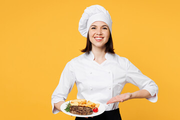 Wall Mural - Young smiling chef cook baker woman wear white shirt black apron uniform toque chefs hat hold in hand point on plate ready dish steak isolated on plain yellow background studio. Cooking food concept.