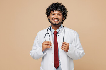 Young smiling happy cheerful Indian doctor man wear white medical gown suit work in hospital clinic office look camera isolated on plain beige background studio portrait. Healthcare medicine concept.