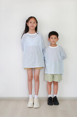 Wall Mural - Asian young girl child and little boy kid standing and looking at camera against white wall in the room. Sister and brother relationship. Full body photo.