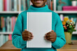 © SnapVault - An African American child in a teal sweater is holding a blank book in front of their chest with both hands. The background shows a blurred bookshelf. Mockup design empty space.