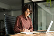 © insta_photos - Busy young Hispanic business woman company owner ceo working in office using laptop writing notes. Serious female professional executive manager in her 30s using computer, elearning at workplace.