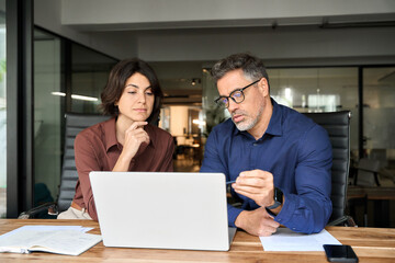 Wall Mural - Two busy professional executives team using laptop in corporate office. Business man and woman leaders managers working on computer at work desk having conversation on financial project at meeting.