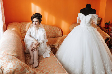 A woman sits on a couch in a room with a wedding dress on a mannequin. The room is decorated with orange walls and a potted plant. The woman is wearing a white gown and a veil