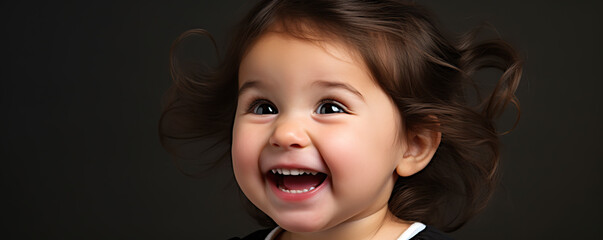 Wall Mural - Happy toddler with curly hair smiles joyfully in a close-up portrait against a dark background during a playful indoor moment