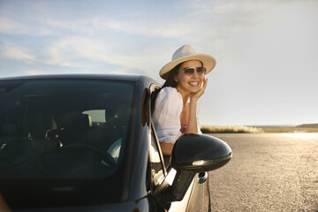 Canvas Print - Smiling young woman in sunglasses with hat leaning out of car window outdoors. Enjoying trip