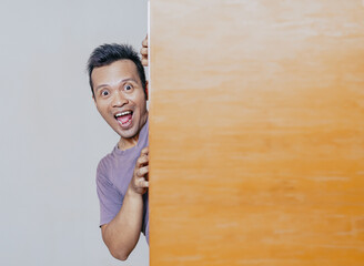 Shocked and surprised face of asian man peeking behind wooden cupboard.