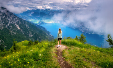 Wall Mural - Young woman on the trail on mountain peak and beautiful view of Garda lake in low clouds at sunset in summer. Dolomites, Italy. Colorful landscape with girl, path, water, green grass, fog, sky. Hiking