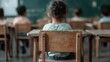 ©  Shomixer - A young child is seen from the back, sitting at a wooden desk in a classroom with a green chalkboard and other students in the background. Learning environment with focused students.