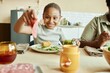 © Seventyfour - Medium shot of smiling African American girl eating roasted corn at table treating herself to slice of sausage during family dinner at beige kitchen