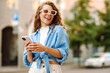 © maxbelchenko - Cheerful young woman in fashionable sunglasses engages with her smartphone, enjoying the beautiful weather on a lively city street filled with greenery. Technology, connection, blogging, modern lifest