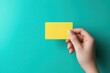 © David Zarzosa - A hand holding a blank yellow card on a turquoise background, used for promotions or designs.