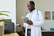 © pressmaster - African American doctor holding medical documents while standing next to printer in well-lit medical office