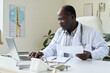 © pressmaster - African American male doctor seated at desk analyzing documents. Medical environment with laptop and stethoscope visible on table