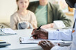 © pressmaster - Healthcare professional filling out medical form while patient stands in background Person not looking at camera View from table showing hand holding pen on paperwork