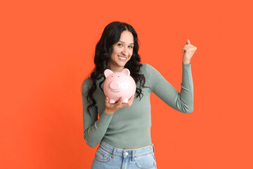 Wall Mural - Pretty young woman with piggy bank pointing at something on orange background