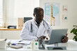 © pressmaster - African American doctor reviewing patient medical records on laptop in office with medical equipment and educational posters visible. Professional workspace and natural lighting